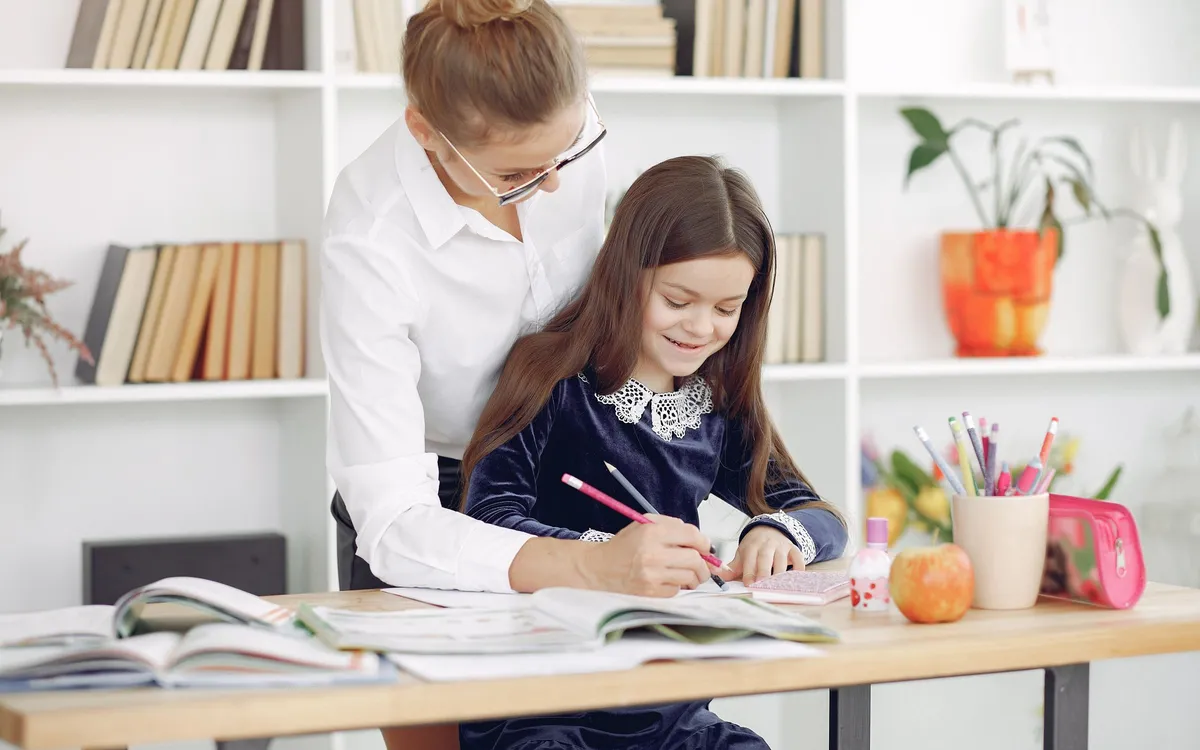 An adult helping a child write in a notebook at a classroom table.